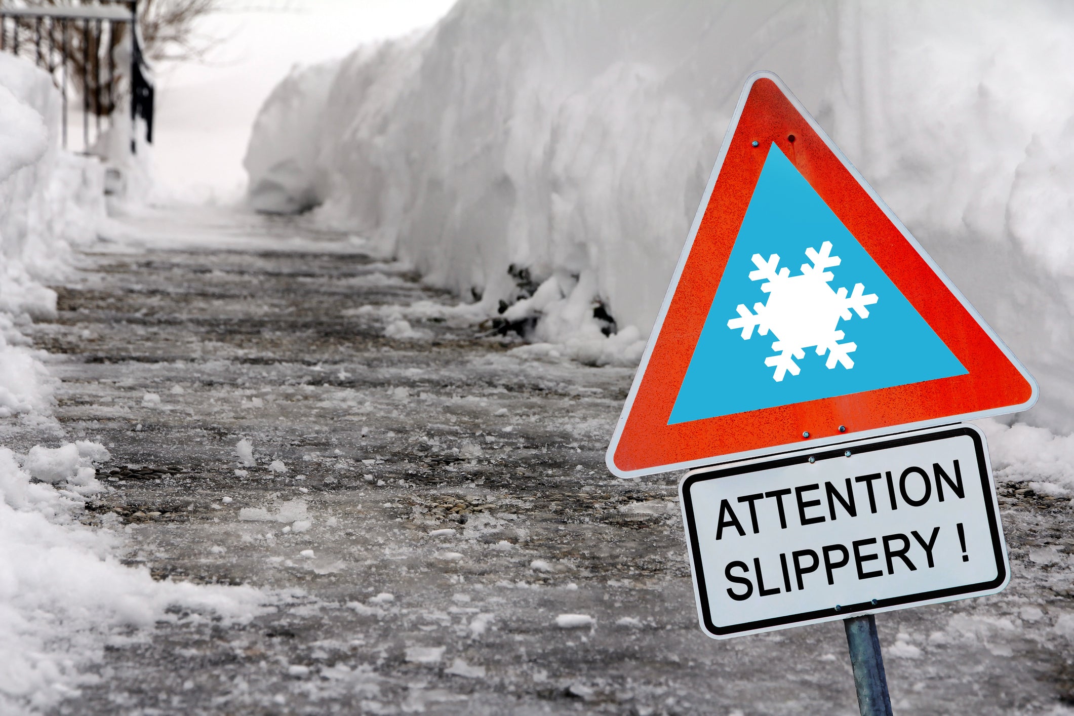 A warning sign with the writing 'Attention Slippery!' is in the foreground and an icy walkway surrounded by snow is in the background