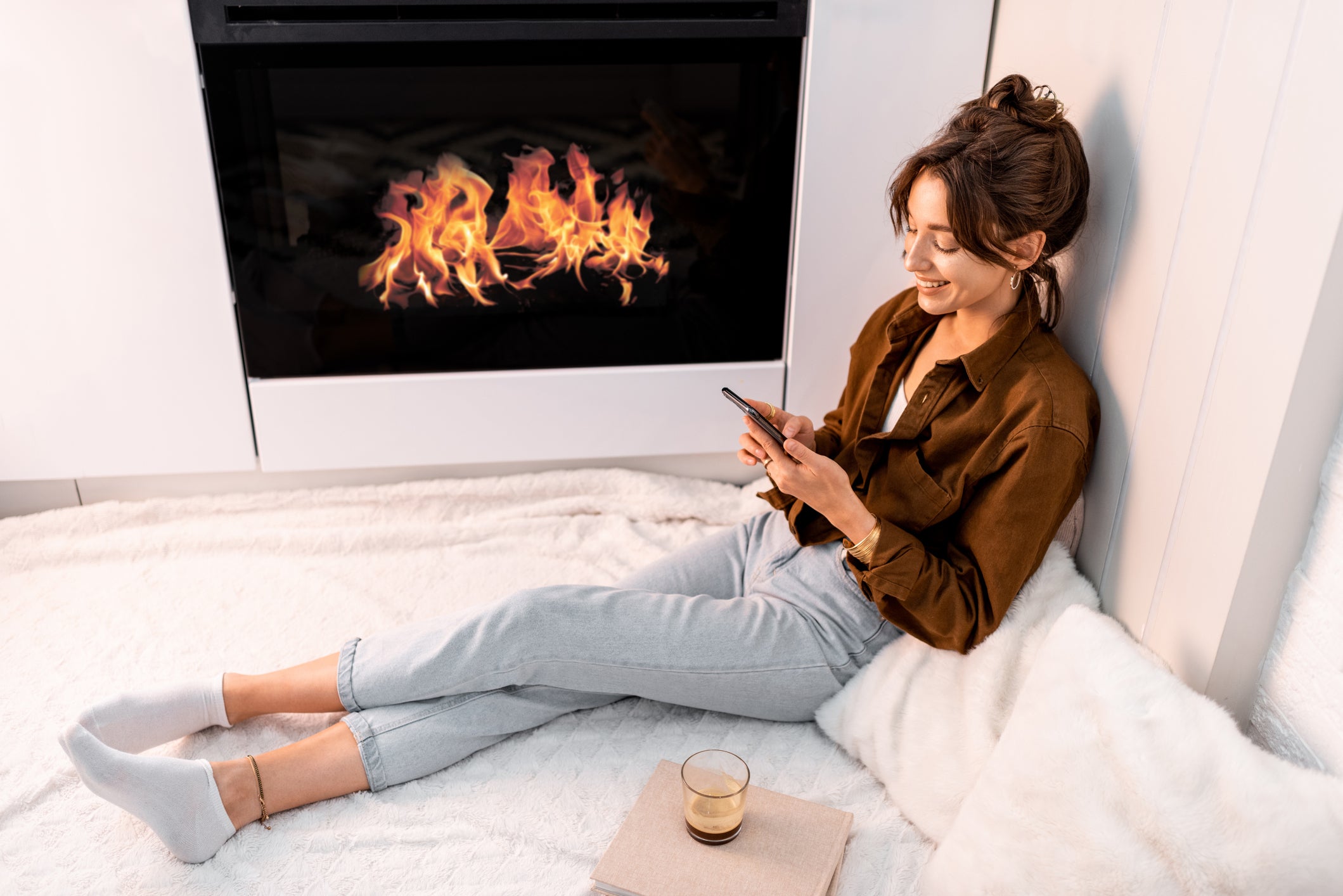 a young woman sits comfortably on the ground near a fireplace with a phone in hand