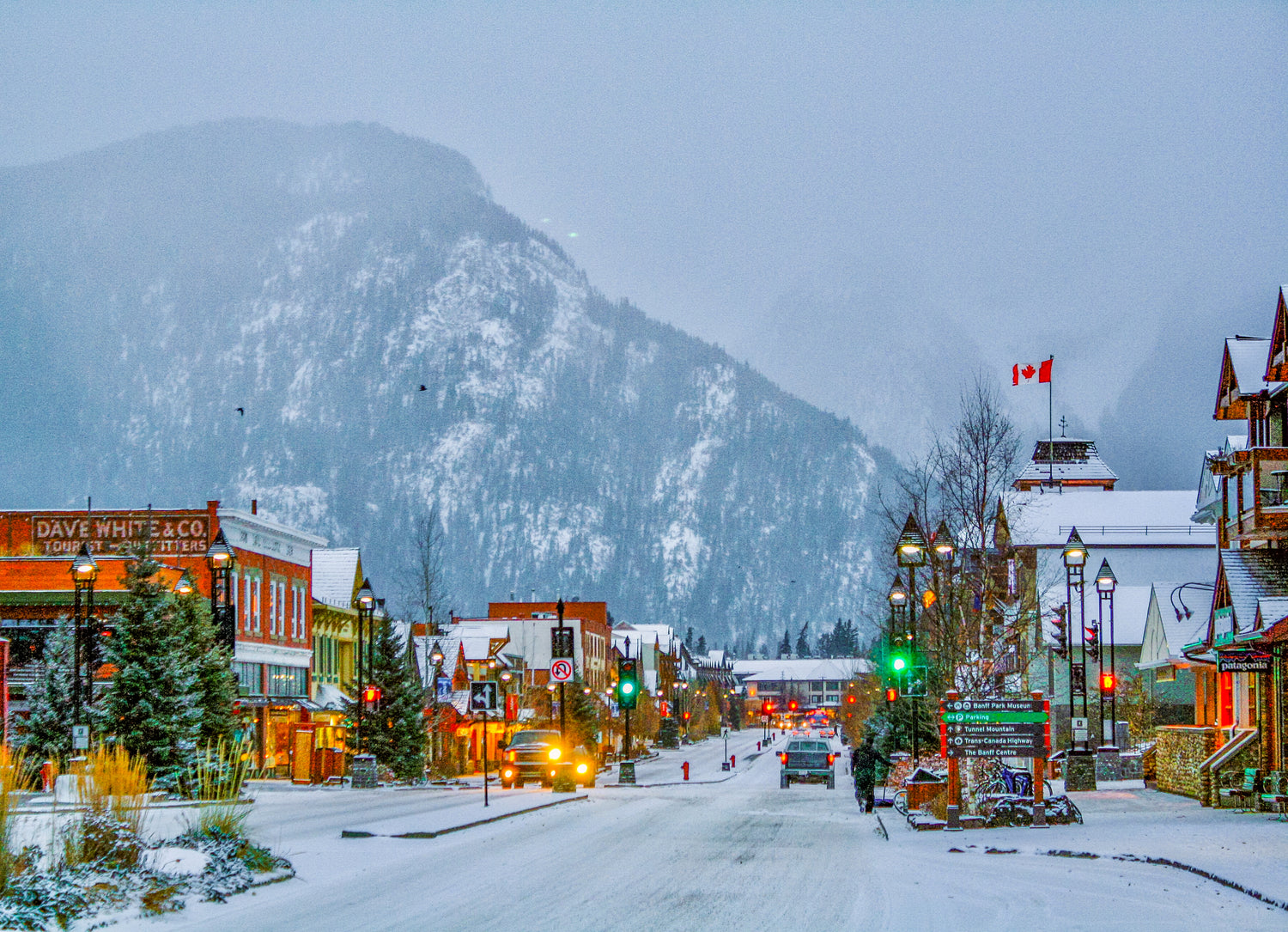 A small town with a Canadian flag is blanketed in snow with tall mountains in the background.