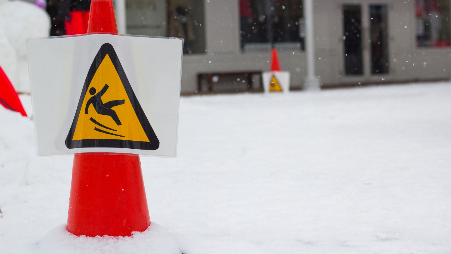 An orange cone has a picture of a stick figure slipping on ice inside a yellow triangle. The ground around the cone is covered in snow.