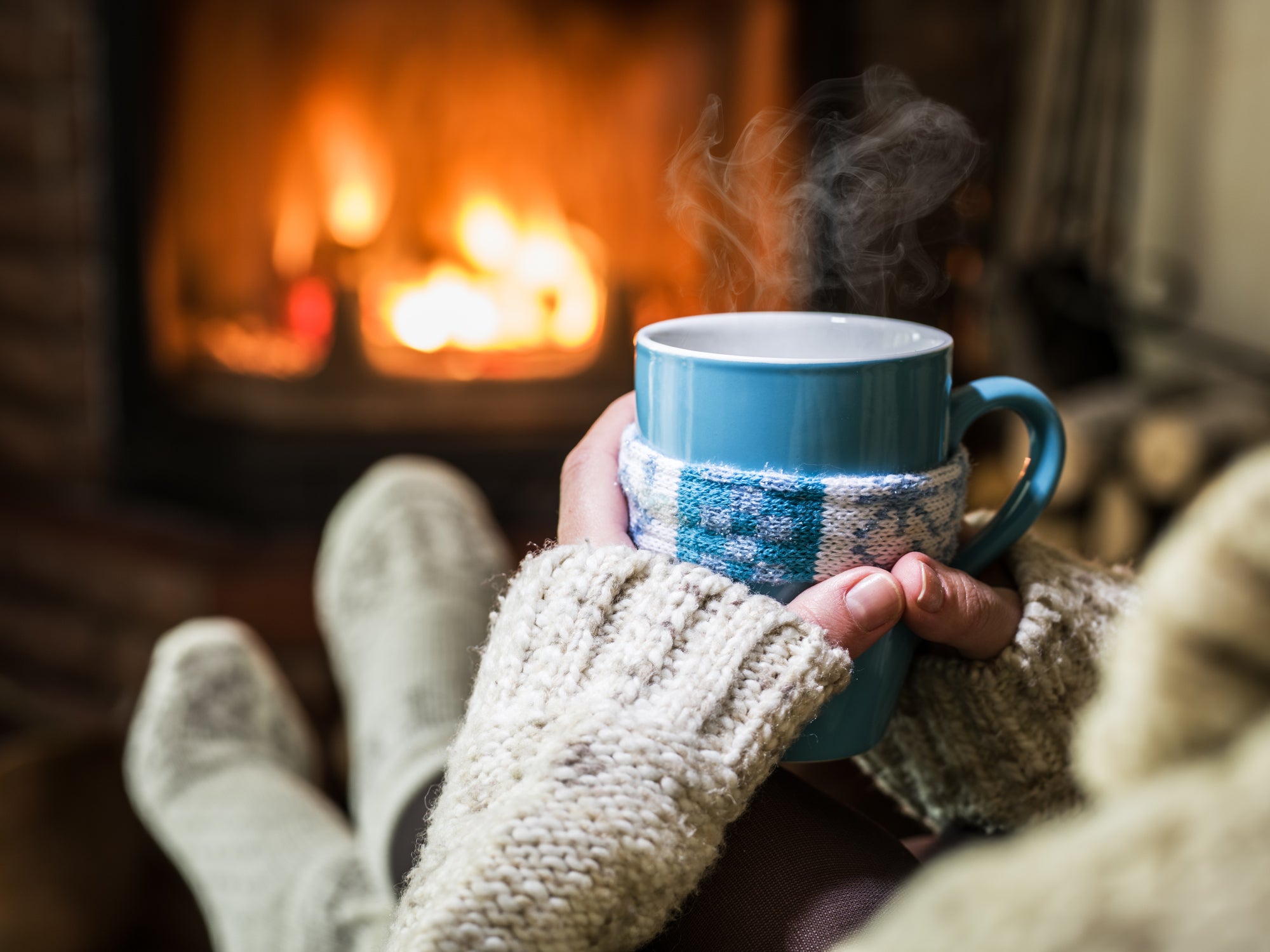 a pair of hands is holding a hot coffee mug near a cozy fireplace
